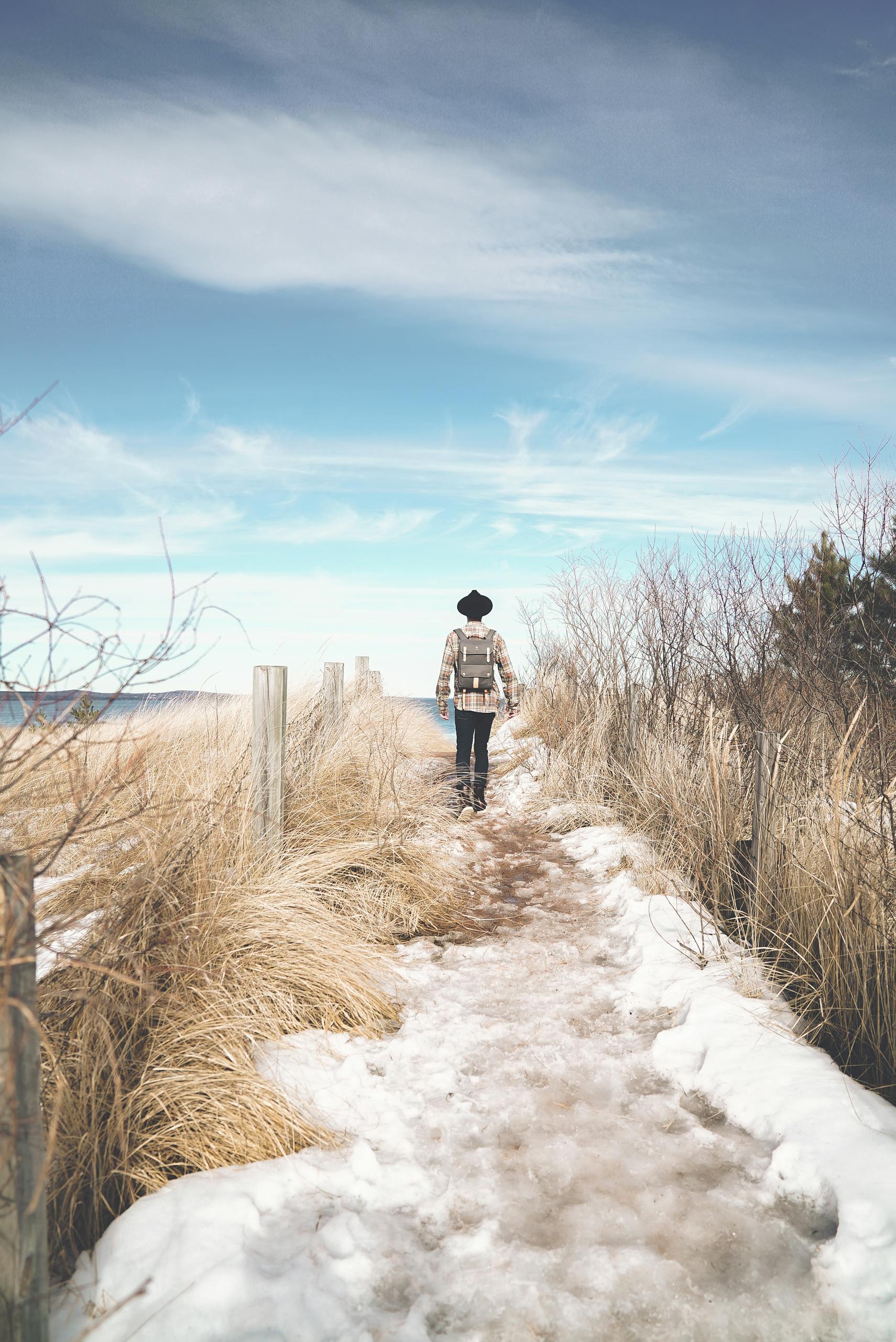 Back view of unrecognizable traveler in trendy outfit and hat walking on snowy path near meadow with tall dry grass against cloudy blue sky in countryside